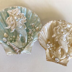 K&ouml;nnte beinhalten: Zwei dekorative Muscheln, jede mit einer Meerjungfrauenfigur. Die Muscheln sind in Perlmuttwei&szlig; und Hellblau bemalt. Die Meerjungfrauen sind silbern und haben langes flie&szlig;endes Haar.