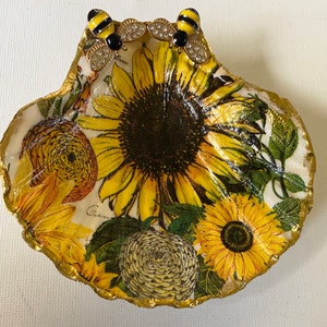 Op de afbeelding: Een decoratieve schelpvormige schaal met een gouden rand en een bloemenontwerp met zonnebloemen, bladeren en een bijenmotief. De schaal is perfect om sieraden of andere kleine voorwerpen in te bewaren.