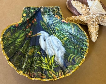 Può includere: Un piatto a forma di conchiglia in porcellana bianca con un bordo dorato. Il piatto è decorato con un airone bianco in piedi in una scena di giungla tropicale con fogliame verde e giallo su uno sfondo blu scuro.