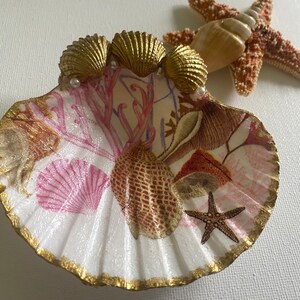 May include: A white seashell dish with a gold rim and a pink and brown seashell design. The dish is decorated with three gold seashells and pearls. A starfish is in the center of the dish.
