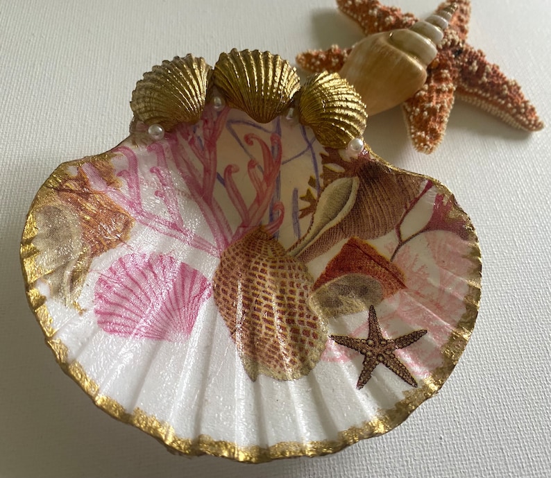 May include: A white seashell dish with a gold rim and a pink and brown seashell design. The dish is decorated with three gold seashells and pearls. A starfish is in the center of the dish.