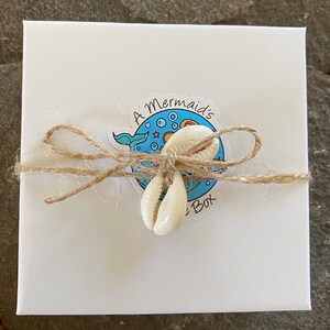 May include: A white box with a blue and white sticker that says "A Mermaid's Box". The box is tied with twine and decorated with two white seashells.