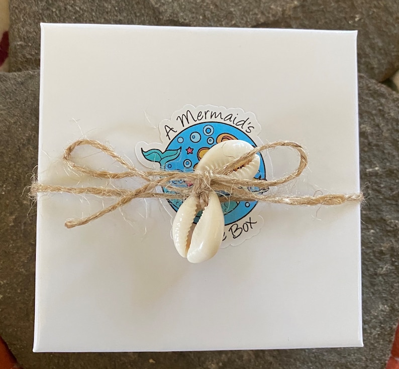 May include: A white box with a blue and white sticker that says "A Mermaid's Box". The box is tied with twine and decorated with two white seashells.