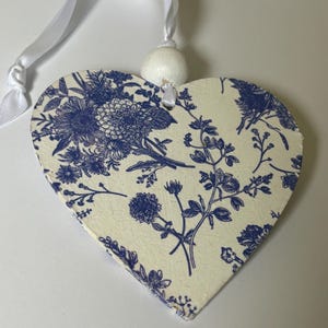 Op de afbeelding: Houten hartvormige ornament met een blauw bloemmotief op een cr&egrave;mekleurige achtergrond. Een wit lint en een kleine witte bal zijn bevestigd om op te hangen. Het bloemmotief bevat verschillende bloemen en bladeren.