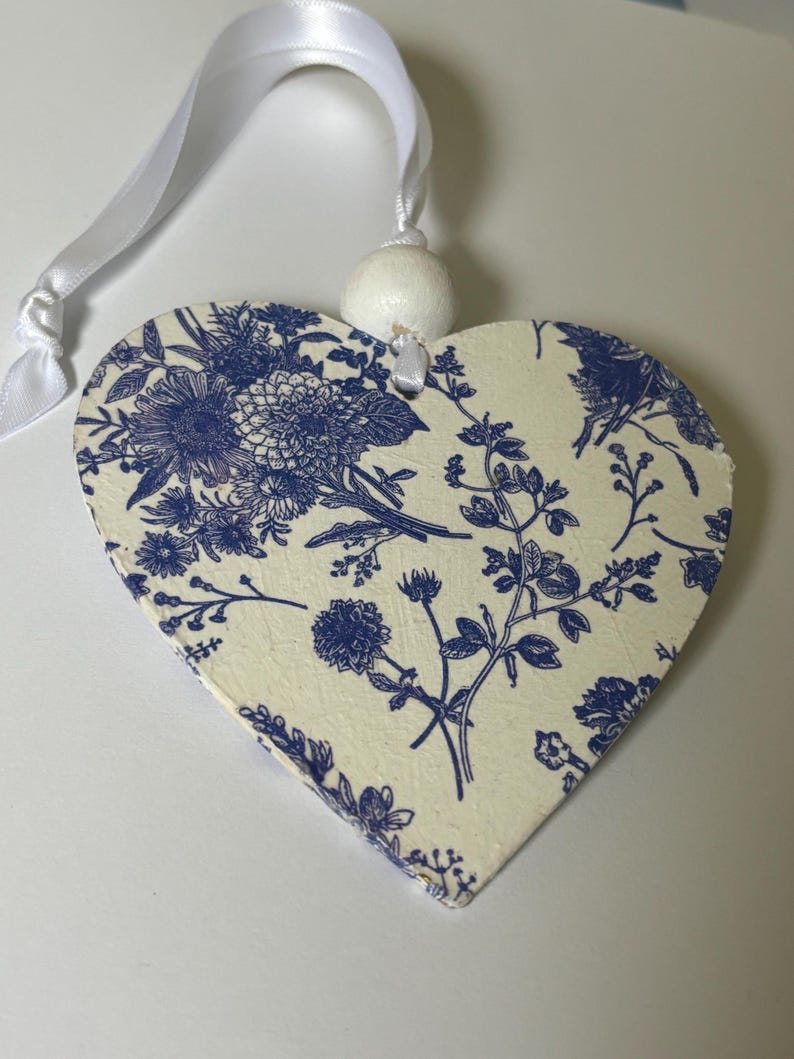 Op de afbeelding: Houten hartvormige ornament met een blauw bloemmotief op een cr&egrave;mekleurige achtergrond. Een wit lint en een kleine witte bal zijn bevestigd om op te hangen. Het bloemmotief bevat verschillende bloemen en bladeren.