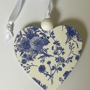 Op de afbeelding: Hartvormige hanger met een blauw bloemmotief op een cr&egrave;mekleurige achtergrond. Het heeft een wit lint en een houten kraal om op te hangen. Het ontwerp toont gedetailleerde bloemen en bladeren.