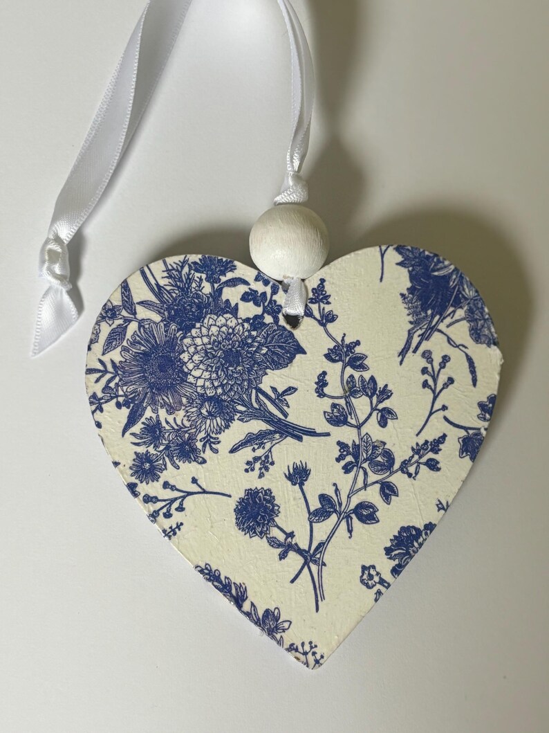 Op de afbeelding: Hartvormige hanger met een blauw bloemmotief op een cr&egrave;mekleurige achtergrond. Het heeft een wit lint en een houten kraal om op te hangen. Het ontwerp toont gedetailleerde bloemen en bladeren.