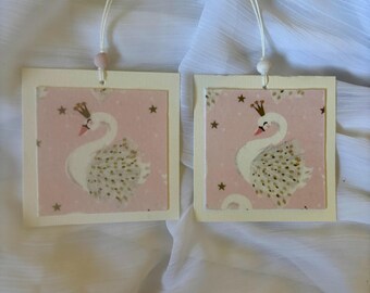 May include: Two white paper gift tags with pink backgrounds and gold stars. Each tag features a white swan with a gold crown and gold accents. The tags are attached to white strings with pink beads.