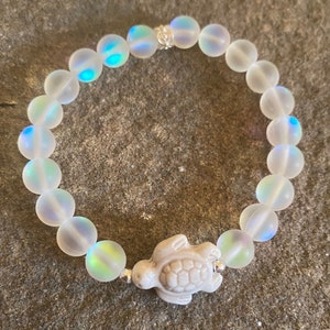 May include: A white iridescent beaded bracelet with a white ceramic turtle charm. The bracelet is made of small, round beads with a subtle iridescent sheen.