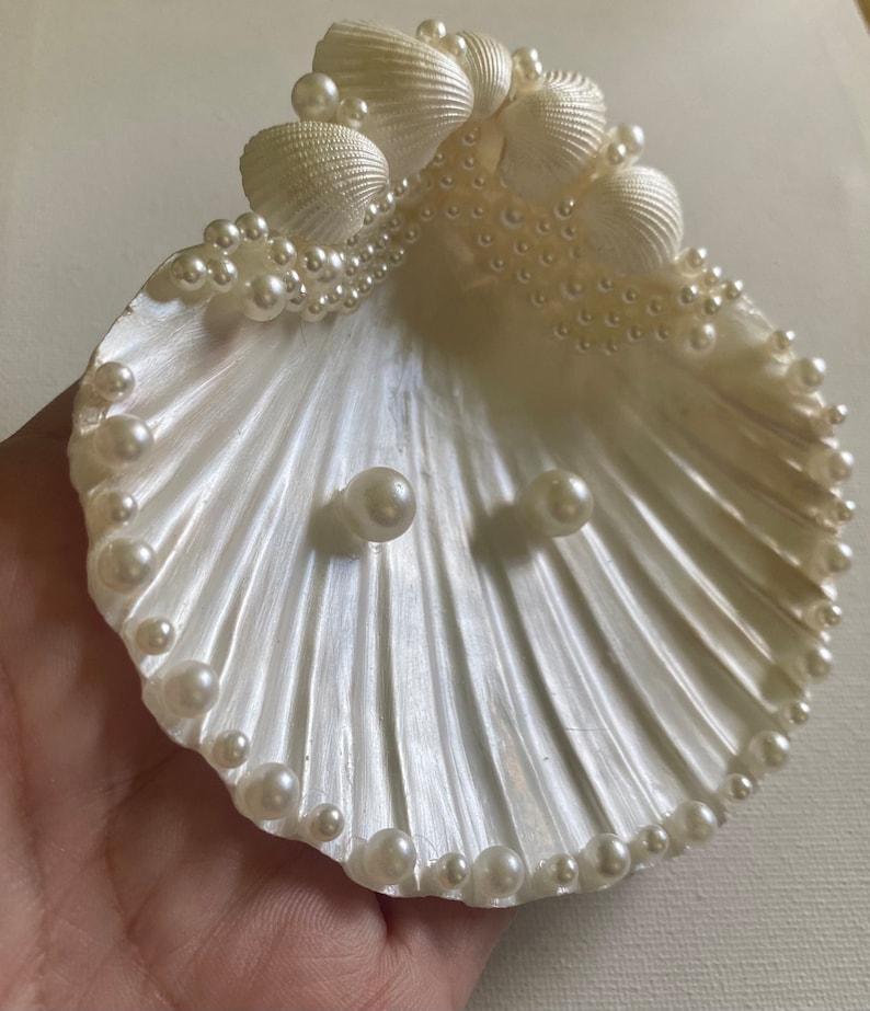 K&ouml;nnte beinhalten: Eine wei&szlig;e Muschel mit einem Perlensaum und zwei Perlen in der Mitte. Die Muschel ist eine dekorative Schale f&uuml;r Schmuck oder andere kleine Gegenst&auml;nde.