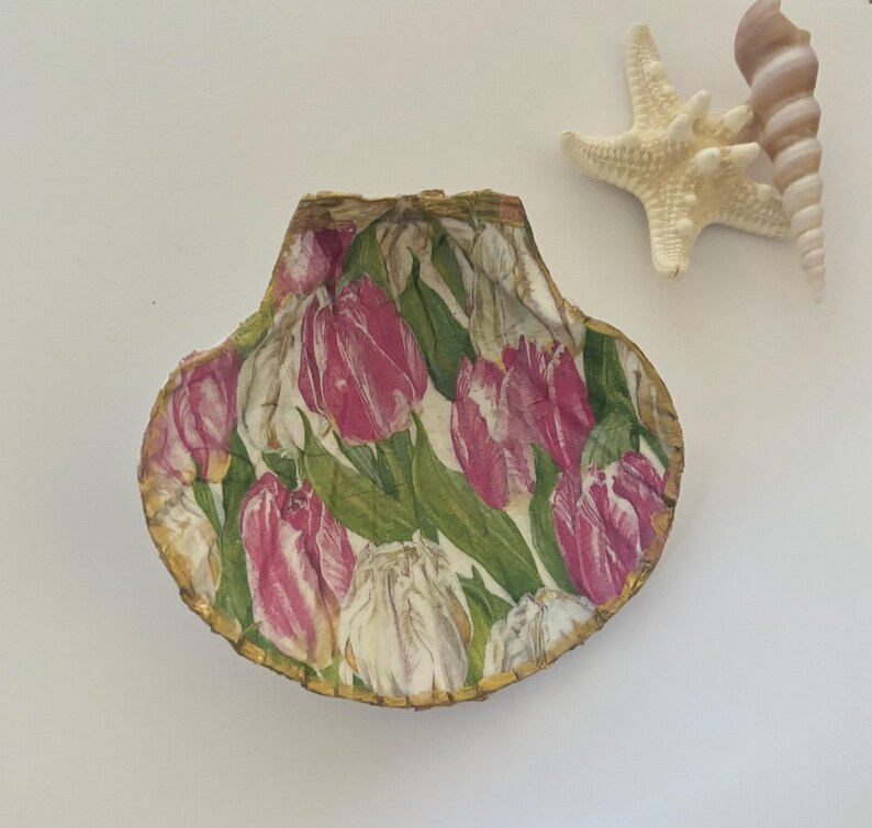 May include: A decorative seashell with a pink and white tulip design. The shell is trimmed with gold paint.
