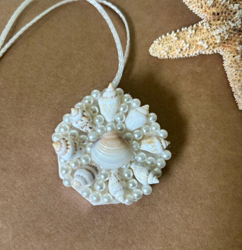 May include: A white pendant necklace with a circular charm featuring a cluster of small white seashells and pearls.