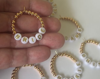 May include: Gold hoop earrings with white beads that spell out the words "BRIDE", "MAC", "BRIT", and "SASH".