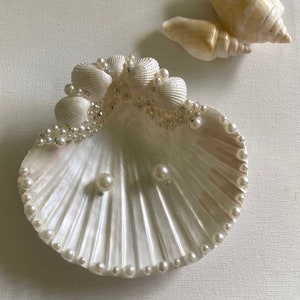 K&ouml;nnte beinhalten: Eine wei&szlig;e Muschelschalen-Schale mit einem Perlensaum und einer Reihe kleinerer Muscheln am oberen Rand. Die Schale ist mit Strasssteinen verziert und enth&auml;lt zwei Perlenohrringe.