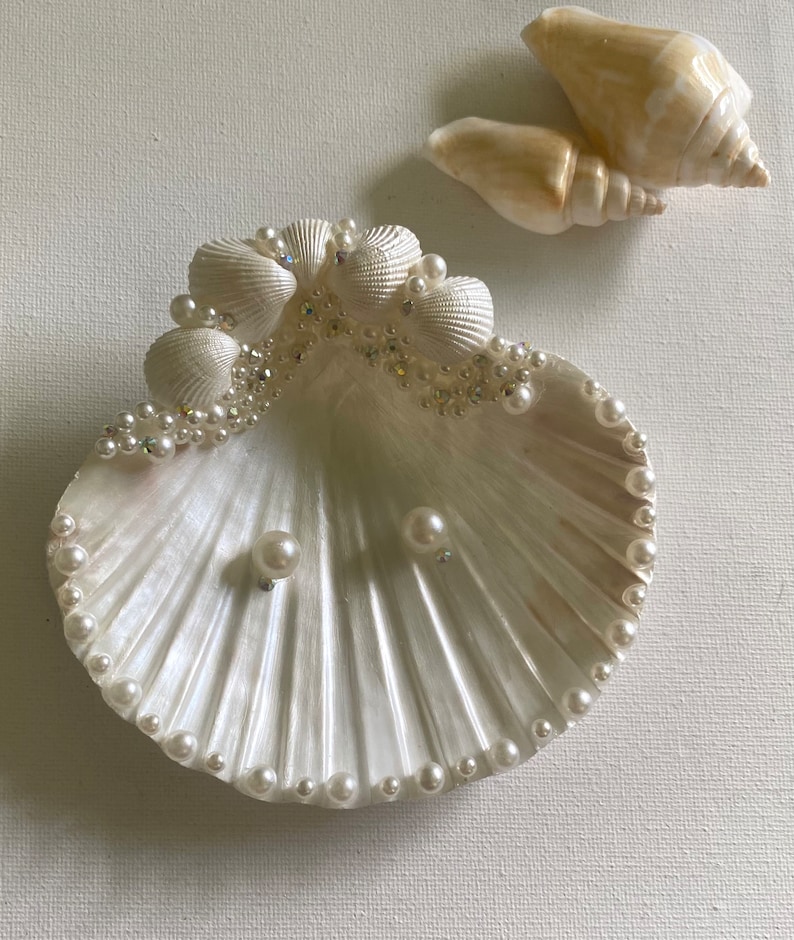 K&ouml;nnte beinhalten: Eine wei&szlig;e Muschelschalen-Schale mit einem Perlensaum und einer Reihe kleinerer Muscheln am oberen Rand. Die Schale ist mit Strasssteinen verziert und enth&auml;lt zwei Perlenohrringe.