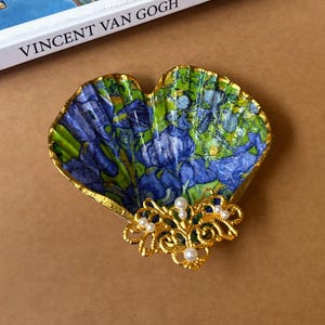 May include: A heart-shaped trinket dish with a blue and green floral design, accented with gold trim and a decorative gold filigree with pearls. The dish is next to a book titled "VINCENT VAN GOGH."