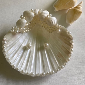 K&ouml;nnte beinhalten: Eine wei&szlig;e Muschelsch&uuml;ssel mit einem Perlensaum und zwei Perlen in der Mitte. Die Sch&uuml;ssel ist mit kleineren Muscheln und Perlen am Rand verziert.