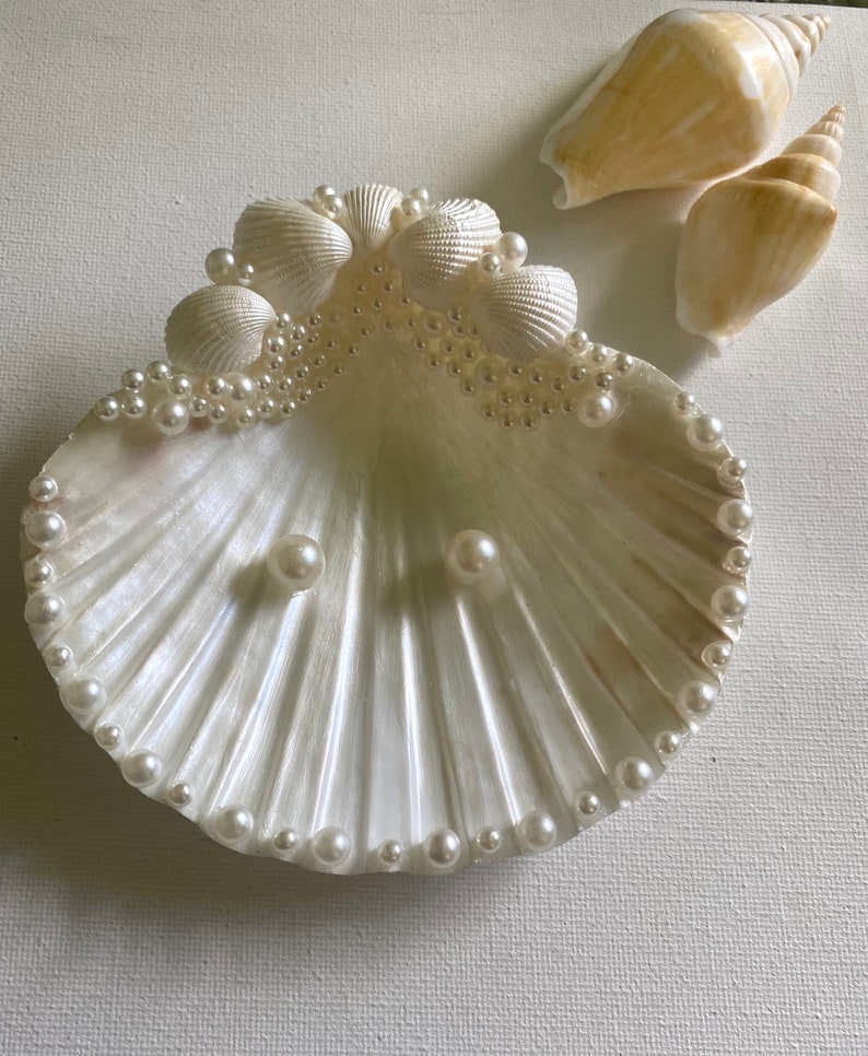 K&ouml;nnte beinhalten: Eine wei&szlig;e Muschelsch&uuml;ssel mit einem Perlensaum und zwei Perlen in der Mitte. Die Sch&uuml;ssel ist mit kleineren Muscheln und Perlen am Rand verziert.