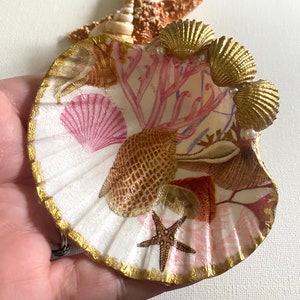May include: A white seashell with a gold rim and a pink and brown seashell design. The seashell is decorated with gold seashells and pearls.