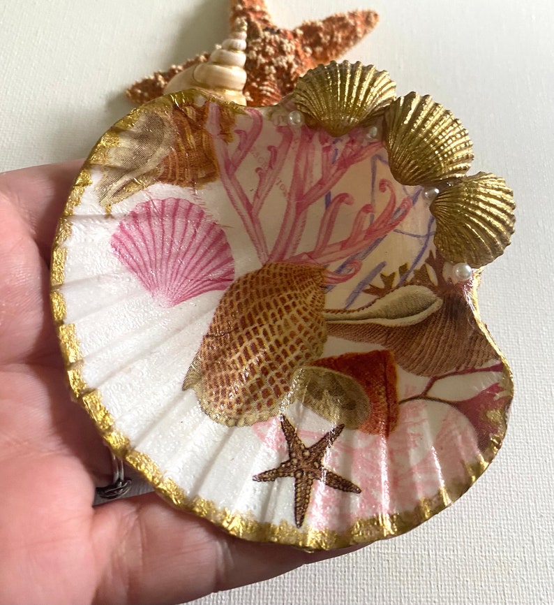 May include: A white seashell with a gold rim and a pink and brown seashell design. The seashell is decorated with gold seashells and pearls.
