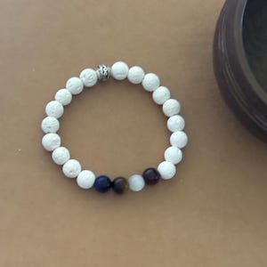 May include: White beaded bracelet with a silver accent bead. The bracelet includes a few accent beads in blue, brown, and dark red. The bracelet is displayed on a light brown surface.