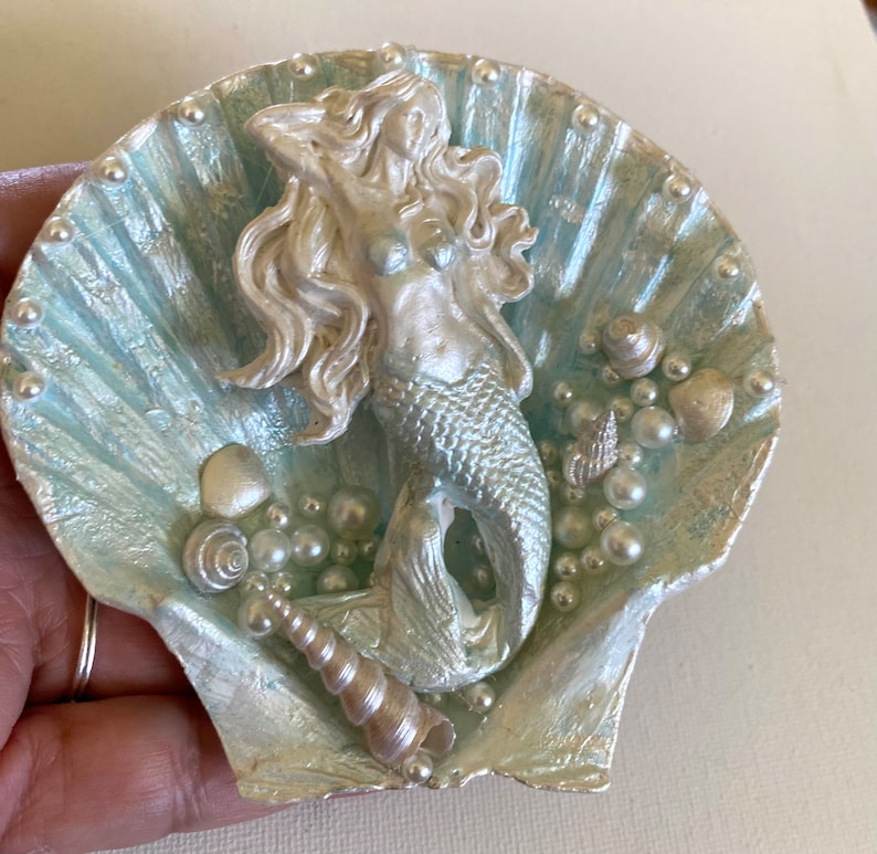 K&ouml;nnte beinhalten: Eine silberne Meerjungfrauenfigur mit langen, flie&szlig;enden Haaren, die in einer hellblauen Muschel mit Perlen- und Muschelakzenten sitzt.