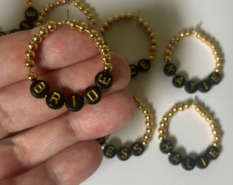 May include: Gold hoop bracelets with black beads spelling out the words "BRIDE", "KATIE", "HALIE", "GRACE", and "JESS".