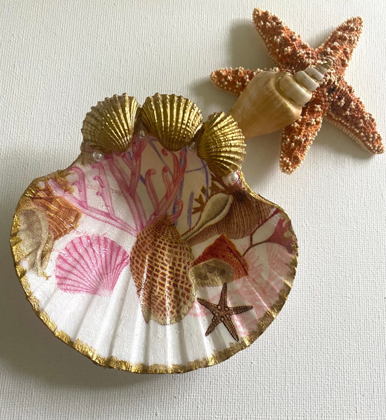 May include: A white seashell dish with a gold rim and a pink and gold seashell design. The dish is decorated with coral, starfish, and pearls.