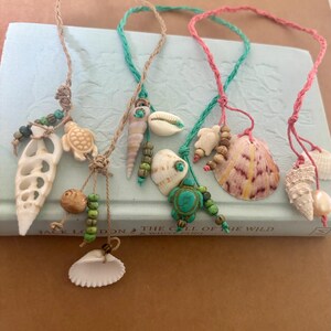 May include: A collection of handmade seashell necklaces. Each necklace features a unique combination of shells, beads, and cord in various colors, including white, beige, green, and pink. The necklaces are displayed on a light blue book.