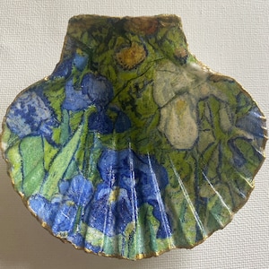 May include: A decorative scallop shell featuring a floral design with blue irises, green foliage, and white flowers. The shell has a gold-painted edge. The artwork is reminiscent of a painting.