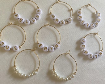 Könnte beinhalten: Sieben goldene Creolen mit weißen Perlen. Sechs der Ohrringe haben weiße Perlen mit goldenen Buchstaben, die Namen buchstabieren: Kelsey, MrsD, Gretchen, Sue, Eryn, Jordyn. Ein Ohrring hat weiße Perlen.