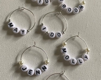 May include: Six silver wine glass charms with white beads and black lettering. The charms spell out the names "Bride", "Groom", "May", "I Do", "Jenn", and "Sam".