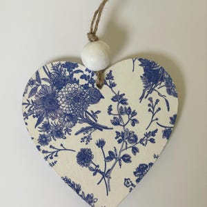 Op de afbeelding: Houten ornament in de vorm van een hart met een blauw bloemmotief op een cr&egrave;mekleurige achtergrond. Het ontwerp toont verschillende bloemen en bladeren. Een witte kraal en een touwlus zijn bevestigd om op te hangen. Het ornament is ongeveer 10 cm breed.