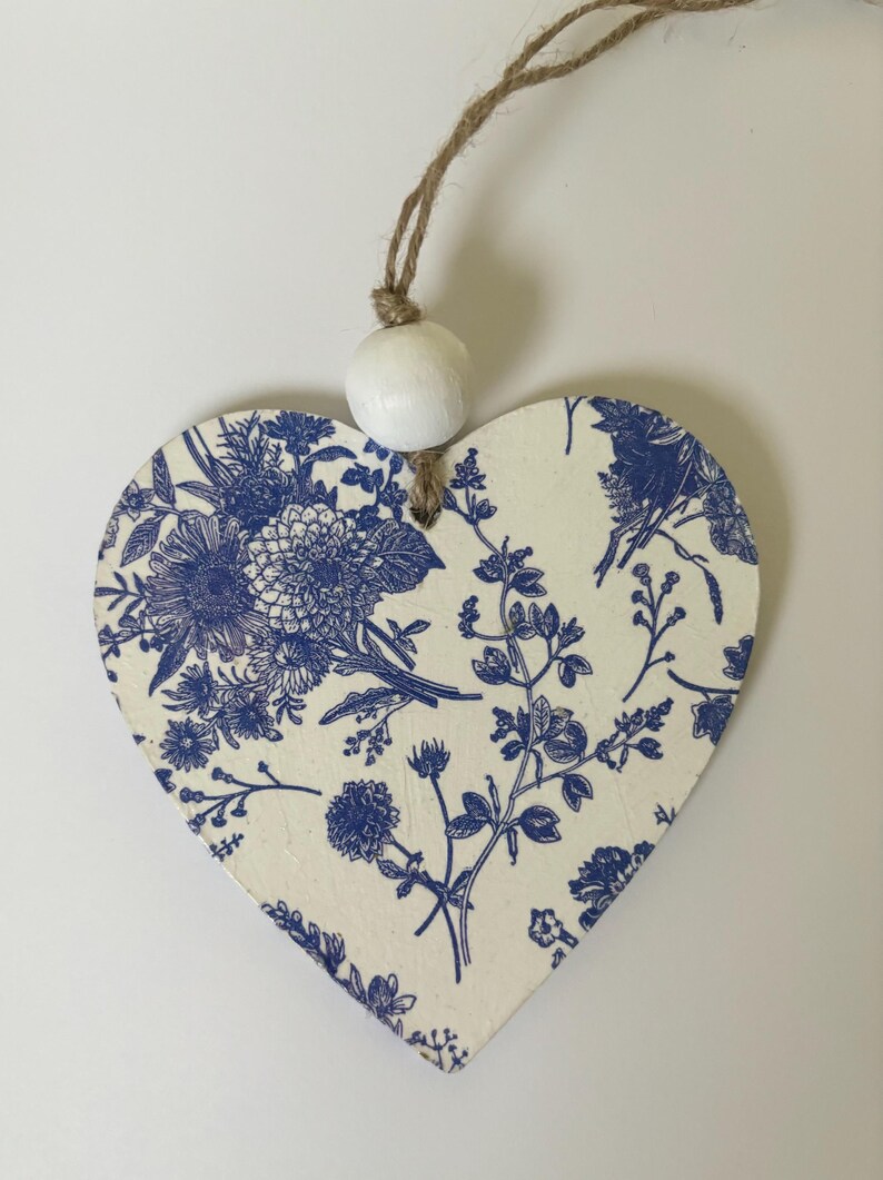 Op de afbeelding: Houten ornament in de vorm van een hart met een blauw bloemmotief op een cr&egrave;mekleurige achtergrond. Het ontwerp toont verschillende bloemen en bladeren. Een witte kraal en een touwlus zijn bevestigd om op te hangen. Het ornament is ongeveer 10 cm breed.