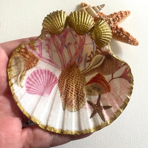 May include: A white seashell with a gold rim and a pink and brown seashell design. The seashell is decorated with three gold seashells and pearls.