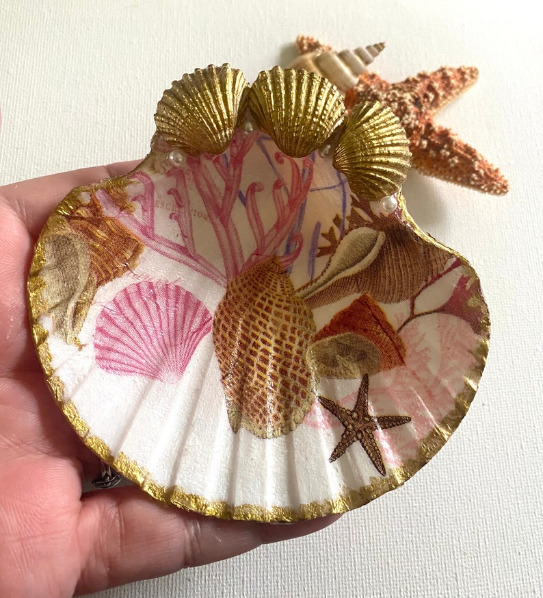 May include: A white seashell with a gold rim and a pink and brown seashell design. The seashell is decorated with three gold seashells and pearls.