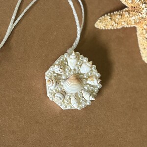 May include: A white hexagonal pendant necklace with a cluster of small seashells and white pearls. The necklace is on a white cord.