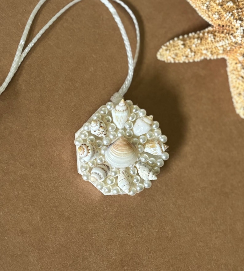 May include: A white hexagonal pendant necklace with a cluster of small seashells and white pearls. The necklace is on a white cord.