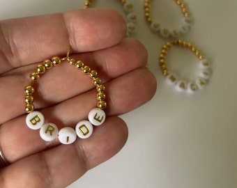 May include: Gold beaded hoop earrings with white letter beads spelling "BRIDE". Additional earrings with names are visible in the background. The earrings are a bridal accessory.