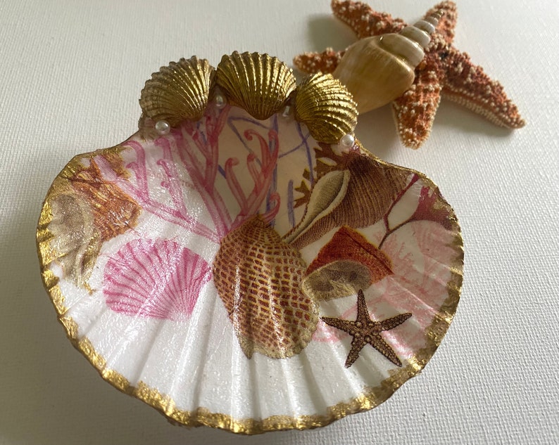 May include: A white seashell dish with a pink and gold floral design. The dish is decorated with gold seashells, pearls, and a starfish.
