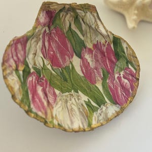 May include: A seashell with a gold rim, decorated with a pink and white floral pattern of tulips. The shell is on a white background.