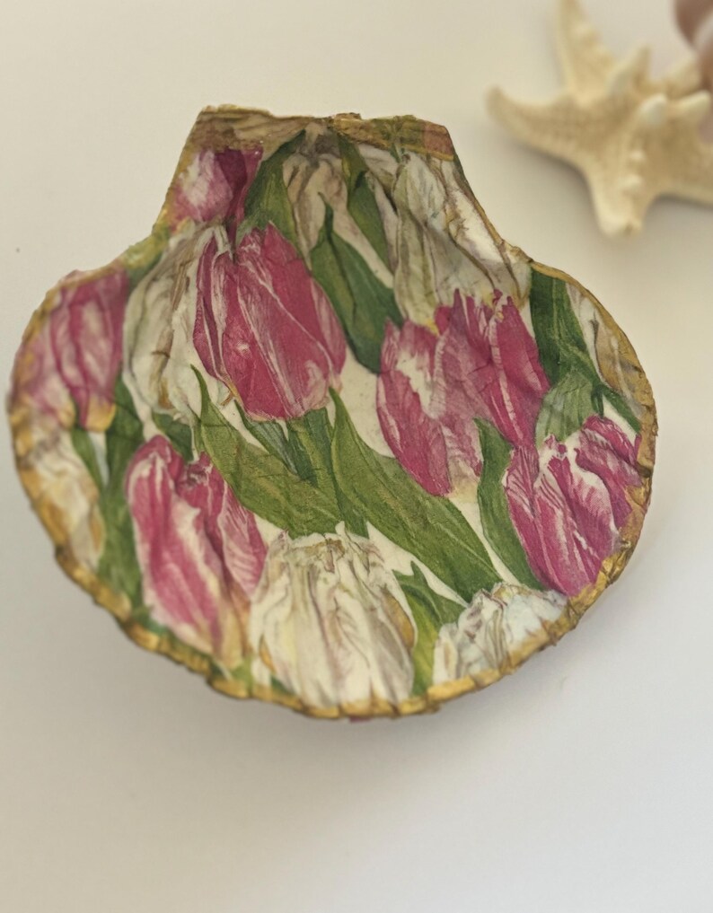 May include: A seashell with a gold rim, decorated with a pink and white floral pattern of tulips. The shell is on a white background.