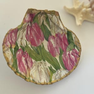 May include: A seashell with a pink and white tulip design. The shell is decorated with a gold trim.