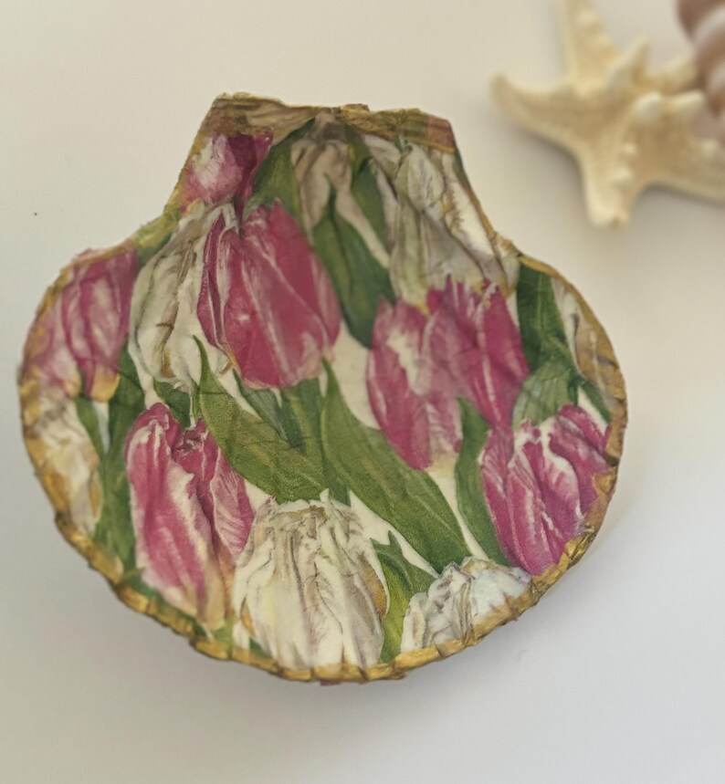 May include: A seashell with a pink and white tulip design. The shell is decorated with a gold trim.