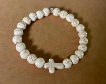 Op de afbeelding: Een witte kralenarmband met een kruisvormige bedel. De armband heeft ronde, crèmekleurige kralen met bruine adering. De kruisbedel is ook wit.
