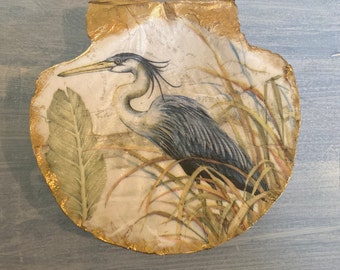 Può includere: Un piatto decorativo rotondo in ceramica, bianco con bordo dorato, con un'immagine dipinta di un airone blu in piedi nell'erba alta.