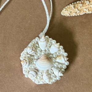 May include: A white seashell ornament with a white cord. The ornament is decorated with small white pearls and various seashells.