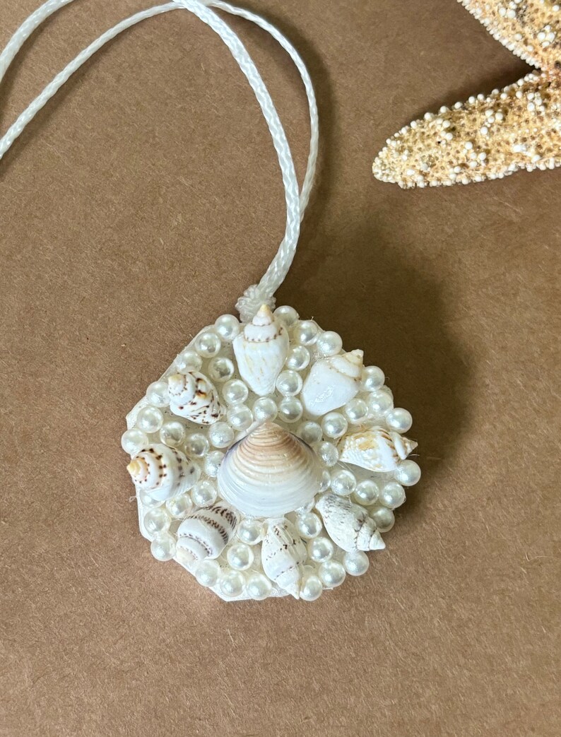 May include: A white seashell ornament with a white cord. The ornament is decorated with small white pearls and various seashells.