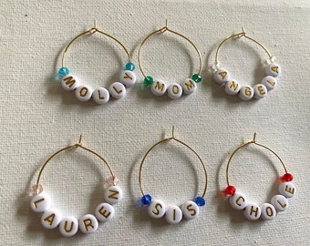 May include: Six gold-colored wine glass charms with white beads spelling out the names "Molly", "Mom", "Angela", "Lauren", "Sis", and "Chloe".