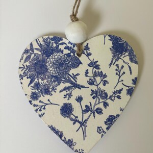 Op de afbeelding: Houten ornament in de vorm van een hart met een blauw bloemmotief op een cr&egrave;mekleurige achtergrond. Het ontwerp toont verschillende bloemen en bladeren. Een witte houten kraal is aan de bovenkant bevestigd met een natuurlijke touwlus om op te hangen.